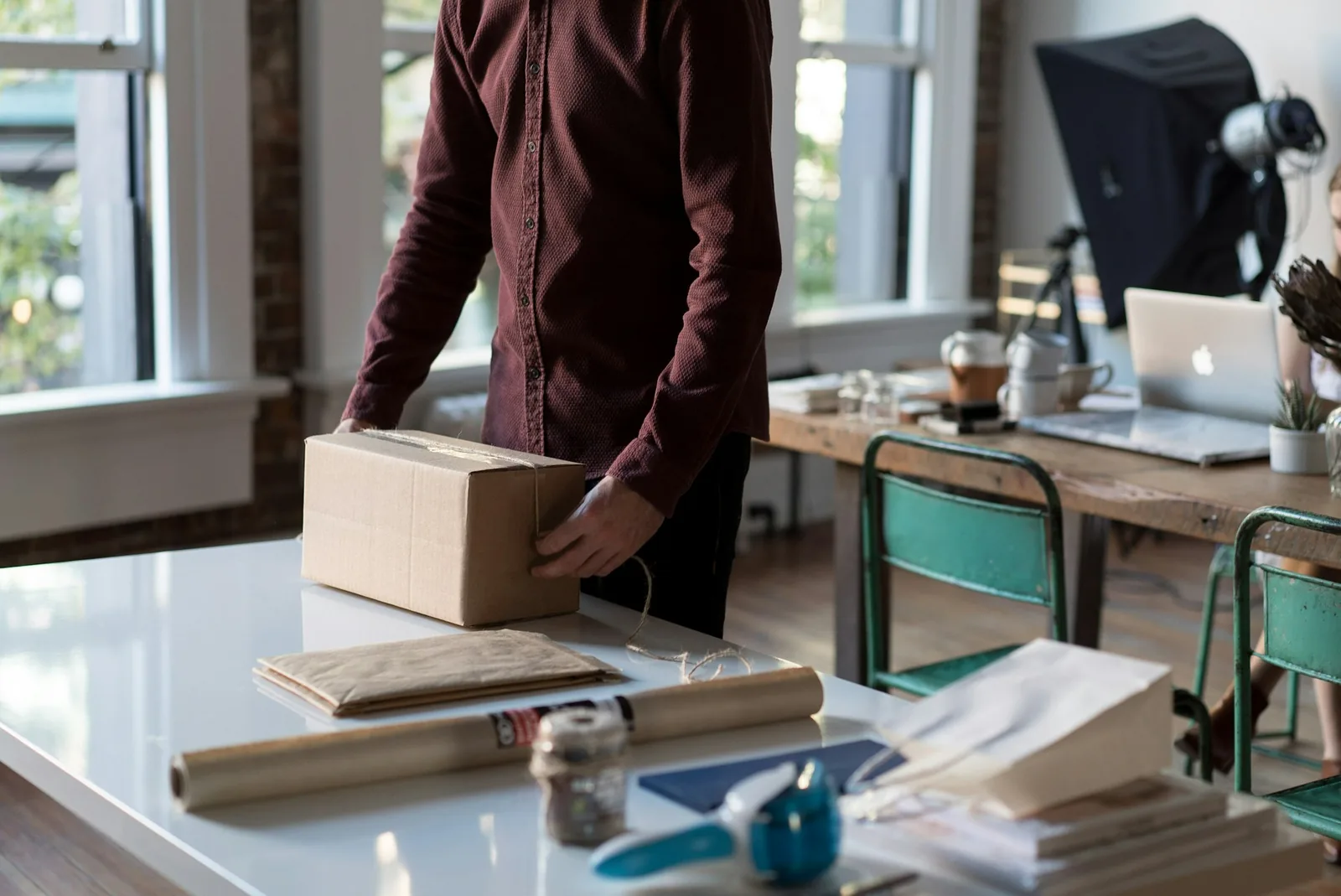 An online seller at a sunlit desk packing a cardboard shipping box, the point where packaging data is captured