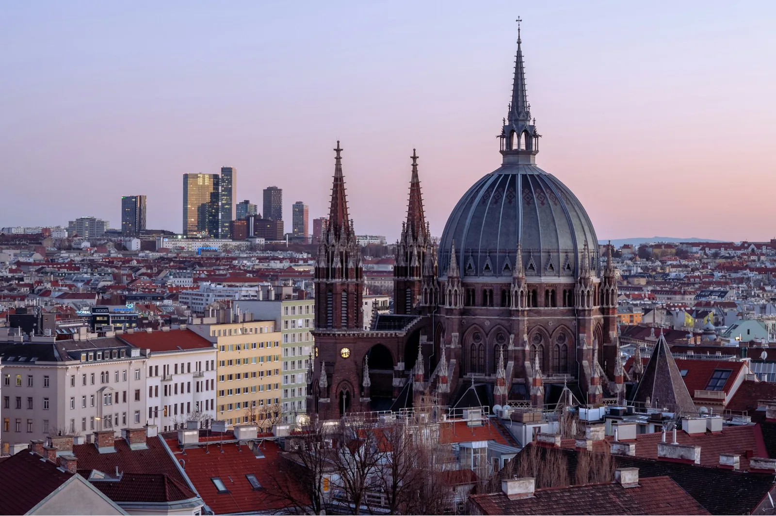 The rooftops of central Vienna at dusk, the dark dome and spires of the Votivkirche rising above tiled roofs under a soft pink sky