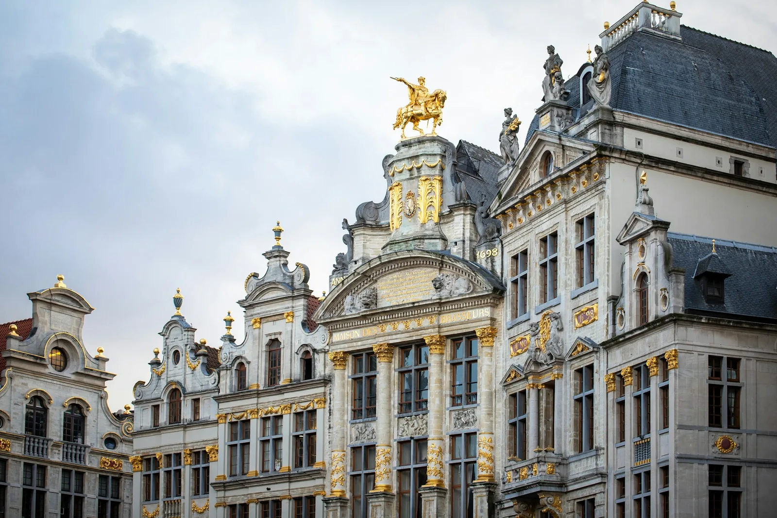 The ornate baroque guild houses of the Grand Place in Brussels, their gilded stone facades and statues rising against a bright cloudy sky
