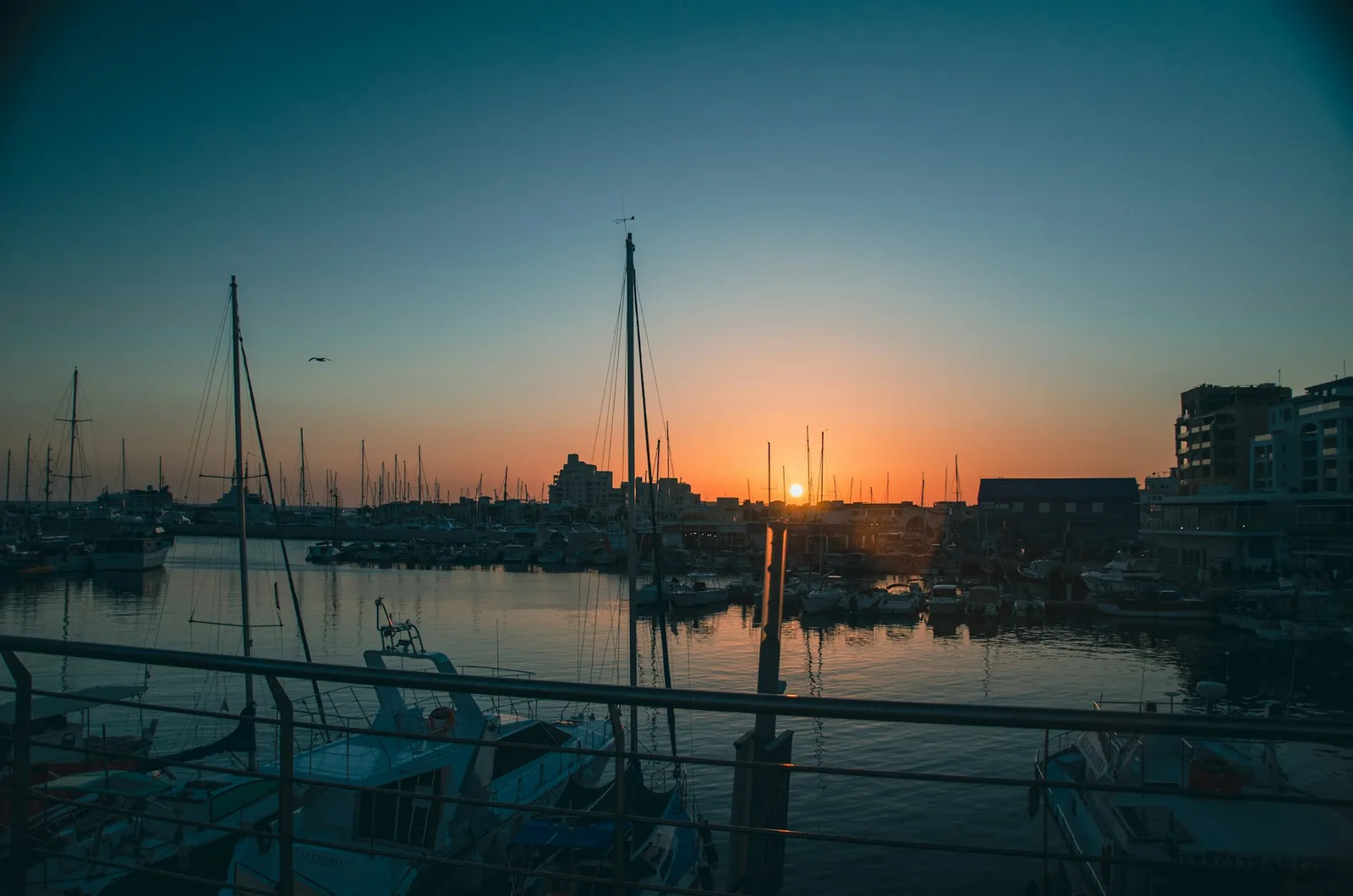 Sailboats moored in a Limassol marina in Cyprus at sunset, silhouetted masts against an orange and blue sky