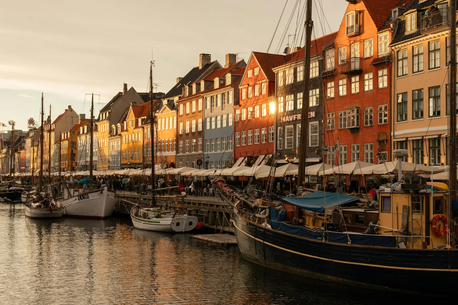 The Nyhavn waterfront in Copenhagen at golden hour, rows of colourful narrow townhouses lining a canal moored with old wooden sailing boats