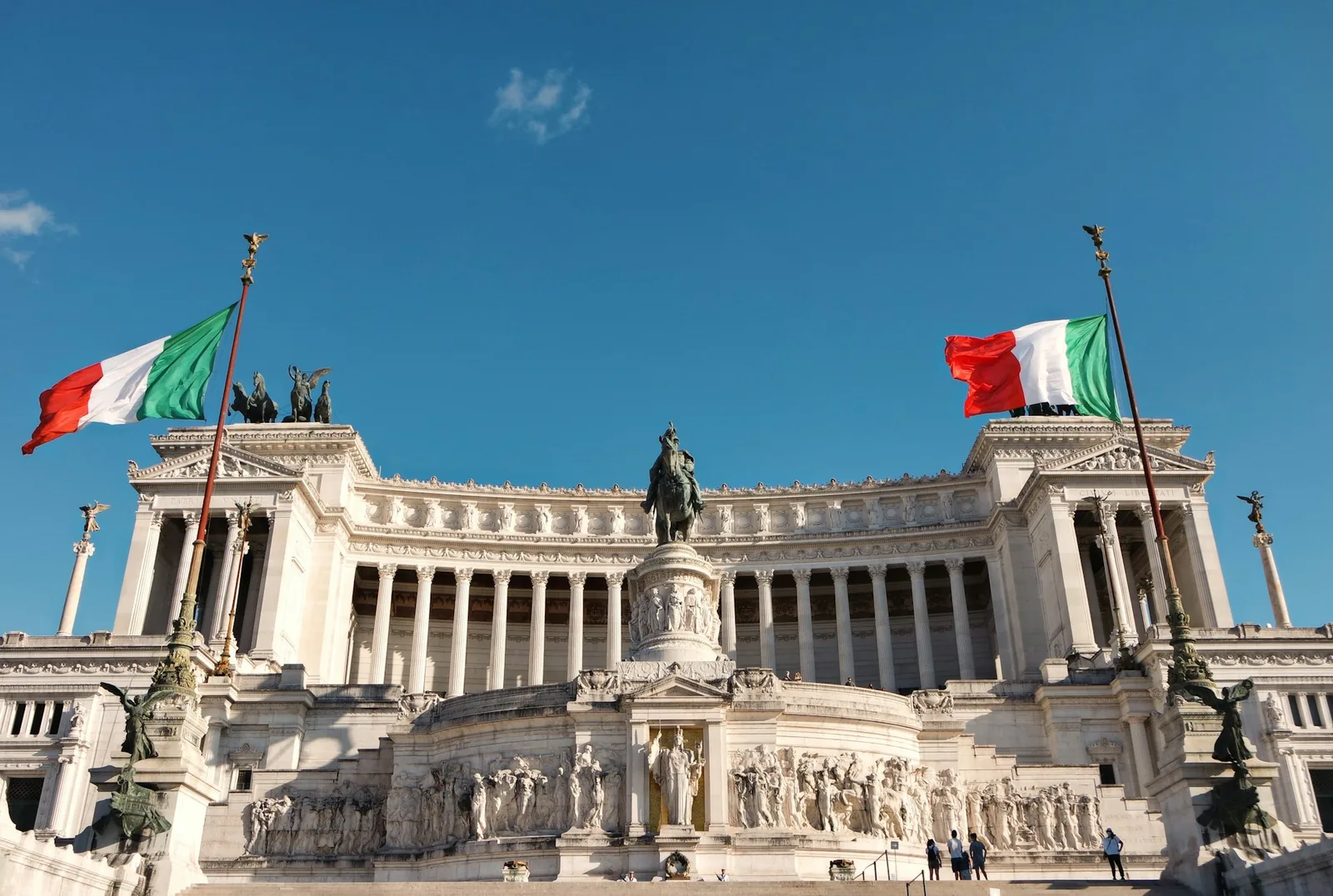 The neoclassical Altare della Patria monument in Rome, its marble colonnade flanked by two Italian flags under a blue sky