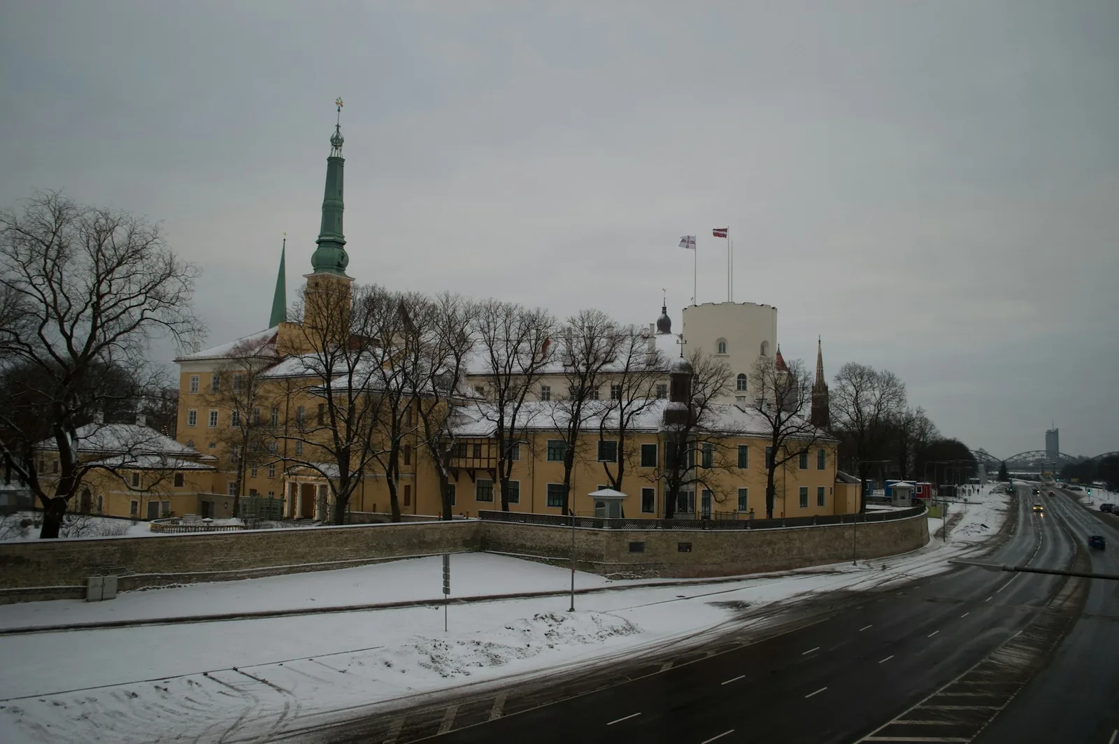 Riga Castle's yellow walls and round tower flying the Latvian flag, seen across a snow-dusted riverside road
