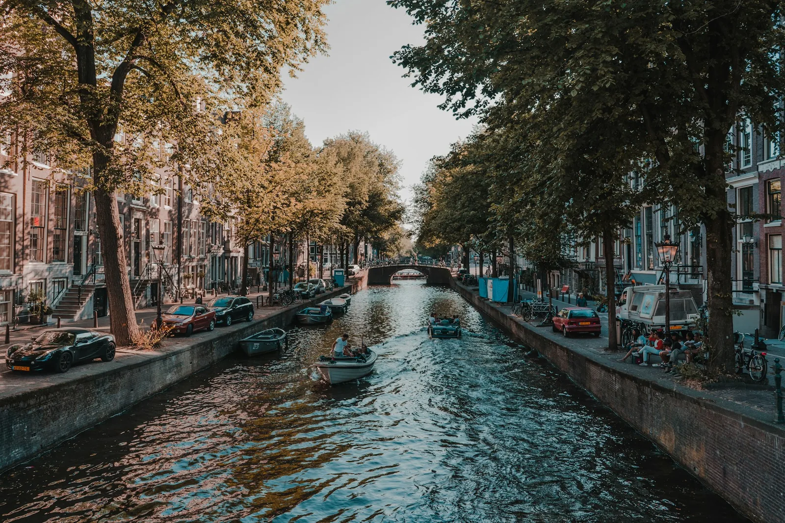 A tree-lined Amsterdam canal with small boats and a stone bridge in warm evening light