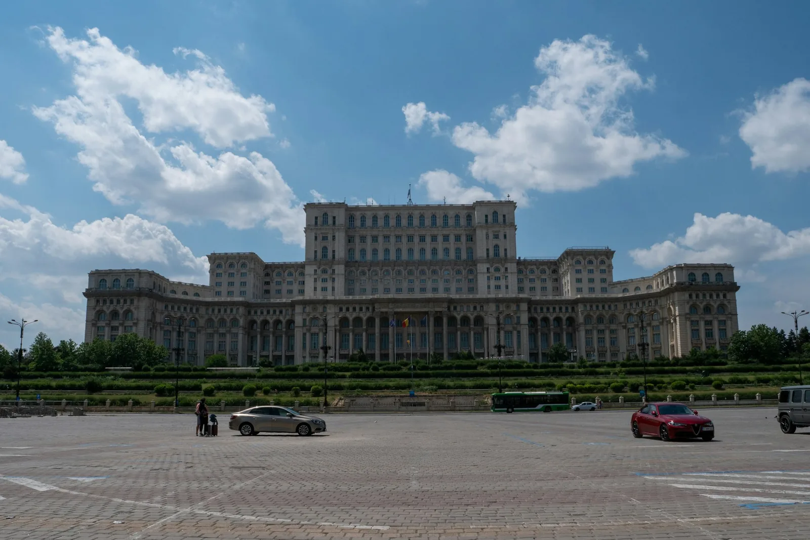 The vast neoclassical Palace of the Parliament in Bucharest under a blue sky with scattered clouds, its columned facade flying the Romanian flag