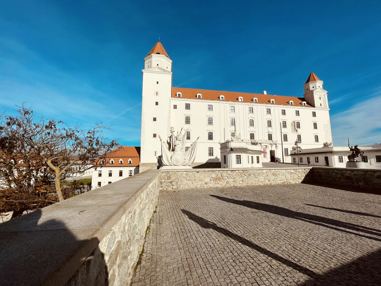 Bratislava Castle's white Baroque walls and orange-roofed corner towers rising above a stone courtyard under a clear blue sky