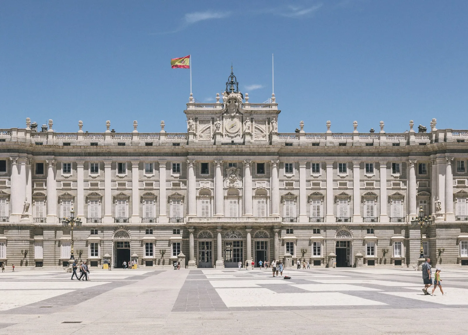 The Royal Palace of Madrid, its symmetrical stone facade and columned courtyard beneath a Spanish flag flying against a clear blue sky